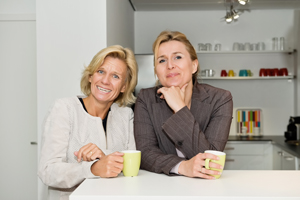 Two women standing behind a kitchen counter and they are holding coffee cups.