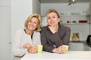 Two women standing behind a kitchen counter and they are holding coffee cups. 